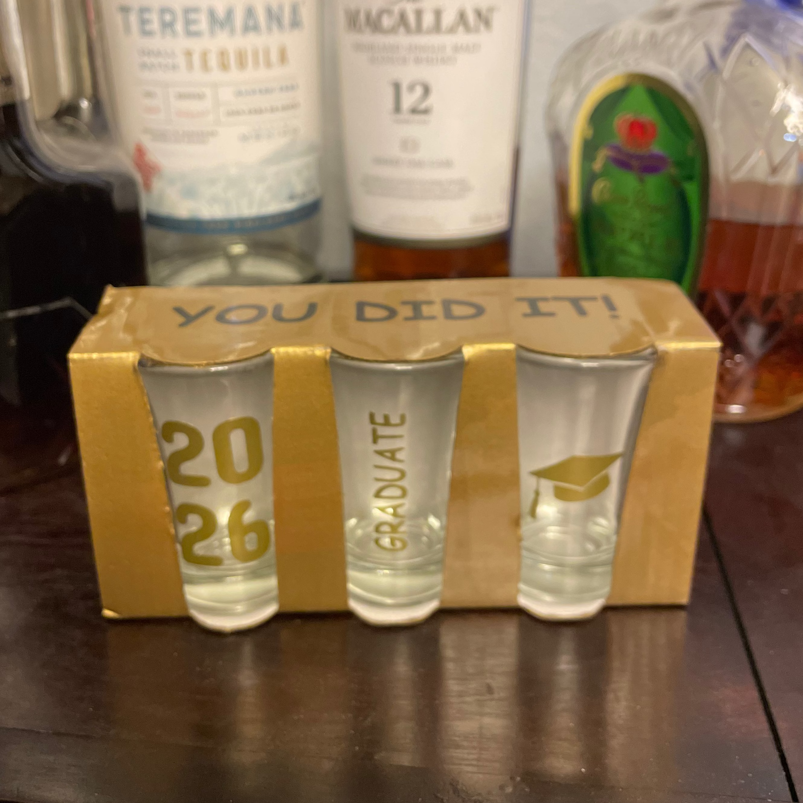 Three shot glasses with '2026 Graduate' text in a box labeled 'You Did It!' surrounded by alcohol bottles.